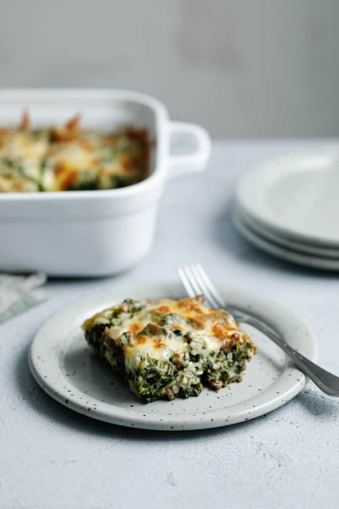 A slice of spinach fandango casserole on a grey speckled plate next to a casserole dish