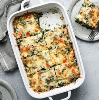A large dish filled with a spinach, cheese and beef casserole with a piece taken out and put on a grey plate with a fork