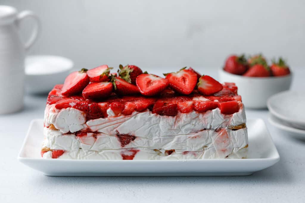 A layered icebox cake dessert with graham crackers, strawberries and cream cheese whip on a white plate surrounded by strawberries and plates