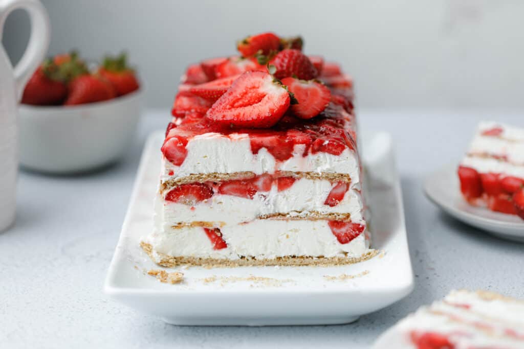 A layered icebox cake dessert with graham crackers, strawberries and cream cheese whip on a white plate surrounded by strawberries and slices of cake