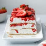 A layered icebox cake dessert with graham crackers, strawberries and cream cheese whip on a white plate surrounded by strawberries and slices of cake