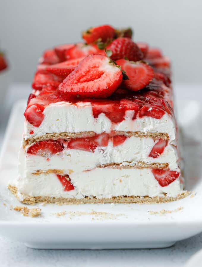 A layered icebox cake dessert with graham crackers, strawberries and cream cheese whip on a white plate surrounded by strawberries and slices of cake