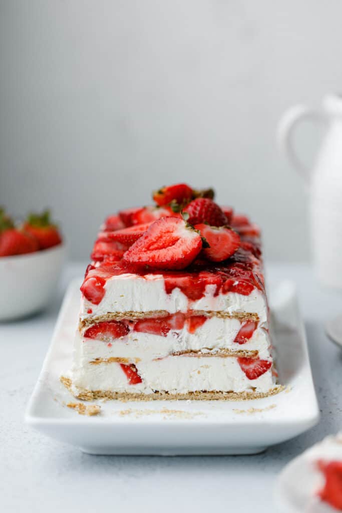 A layered icebox cake dessert with graham crackers, strawberries and cream cheese whip on a white plate surrounded by strawberries and slices of cake.