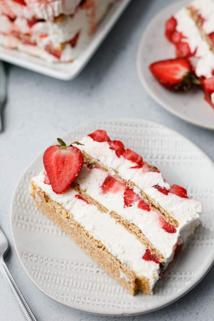Slices of a layered icebox cake dessert with graham crackers, strawberries and cream cheese whip on a white textured plates with a fork.