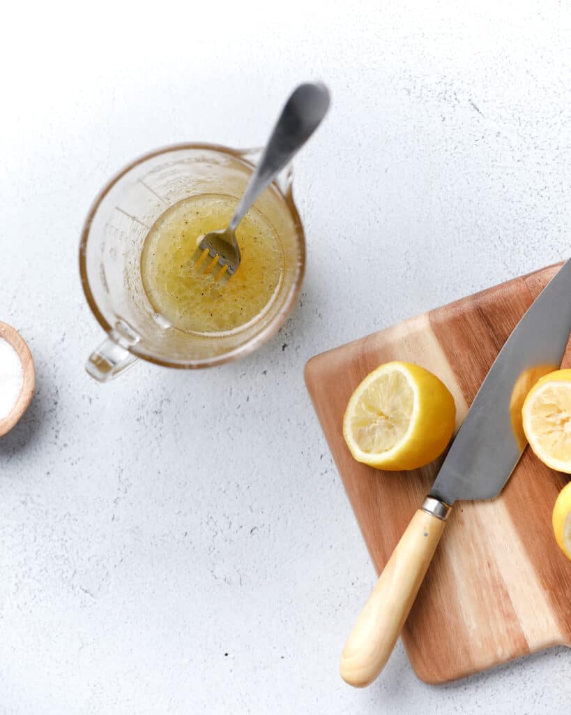 A glass measuring cup filled with salad dressing with cut up lemons on the side on a cutting board.