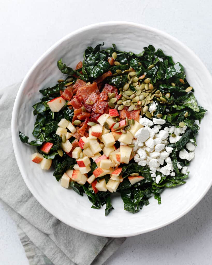 A white bowl filled with a kale salad with bacon, apples, goat cheese and pumpkin seeds.
