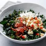 A white bowl filled with kale salad topped with goat cheese, bacon, pepitas, and apple