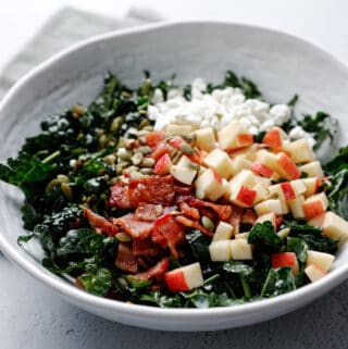 A white bowl filled with kale salad topped with goat cheese, bacon, pepitas, and apple