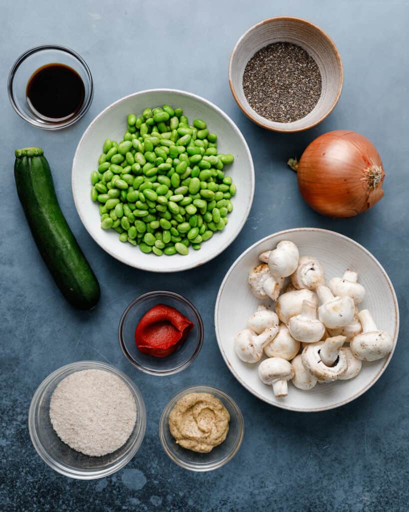 An overhead shot of the ingredients to make an edamame mushroom veggie burger including zucchini, tomato paste, mustard, onions, chia, soy sauce and psyllium husk
