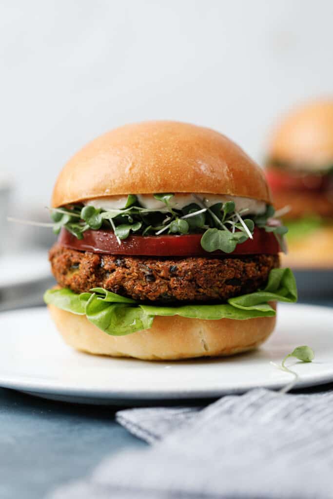 A straight on view of a veggie burger on a white plate with a blue napkin. The burger has a soft bun, mixed greens, butter lettuce, and a slice of heirloom tomato.