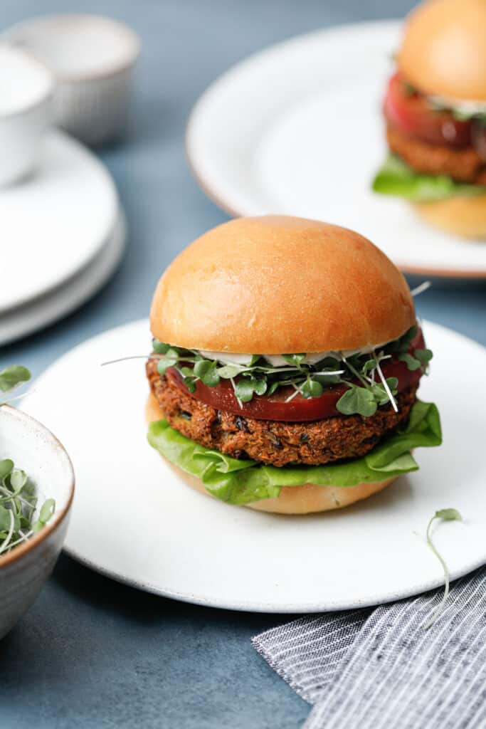 Two veggie burgers on white plates on a blue background with toasted buns, green sprouts, butter lettuce and heirloom tomato.