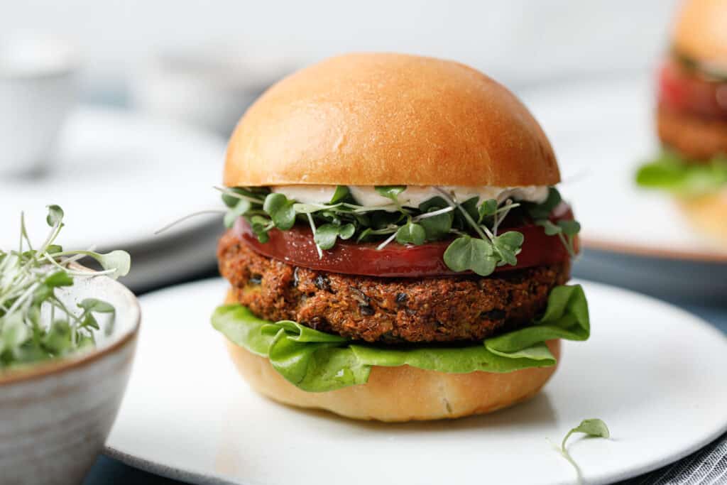 Two veggie burgers on white plates on a blue background with toasted buns, green sprouts, butter lettuce and heirloom tomato.