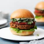 Two veggie burgers on white plates on a blue background with toasted buns, green sprouts, butter lettuce and heirloom tomato.