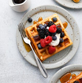 Three waffles seen overhead on plates with forks topped with sour cream and berries with maple syrup being drizzled on