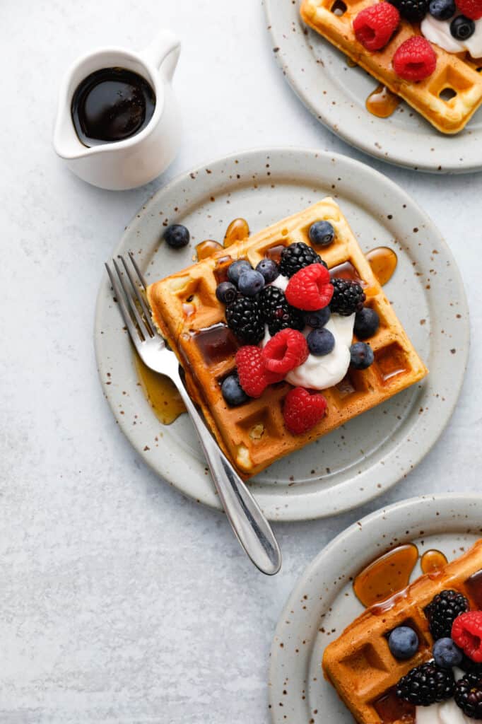 Three waffles seen overhead on plates with forks topped with sour cream and berries with maple syrup being drizzled on