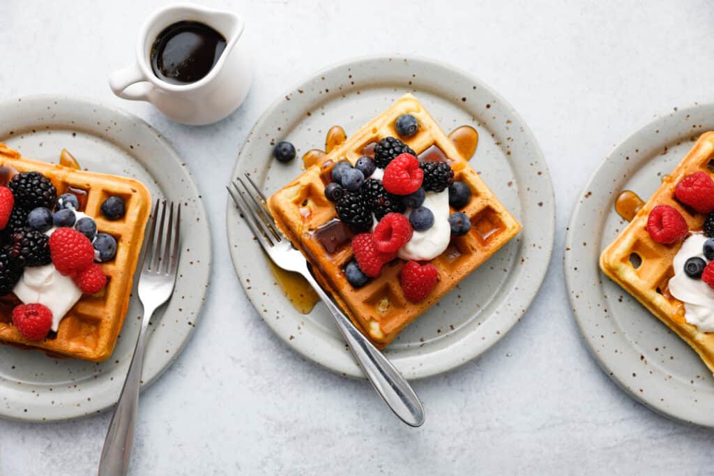 Three waffles seen overhead on plates with forks topped with sour cream and berries with maple syrup being drizzled on