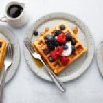 Three waffles seen overhead on plates with forks topped with sour cream and berries with maple syrup being drizzled on