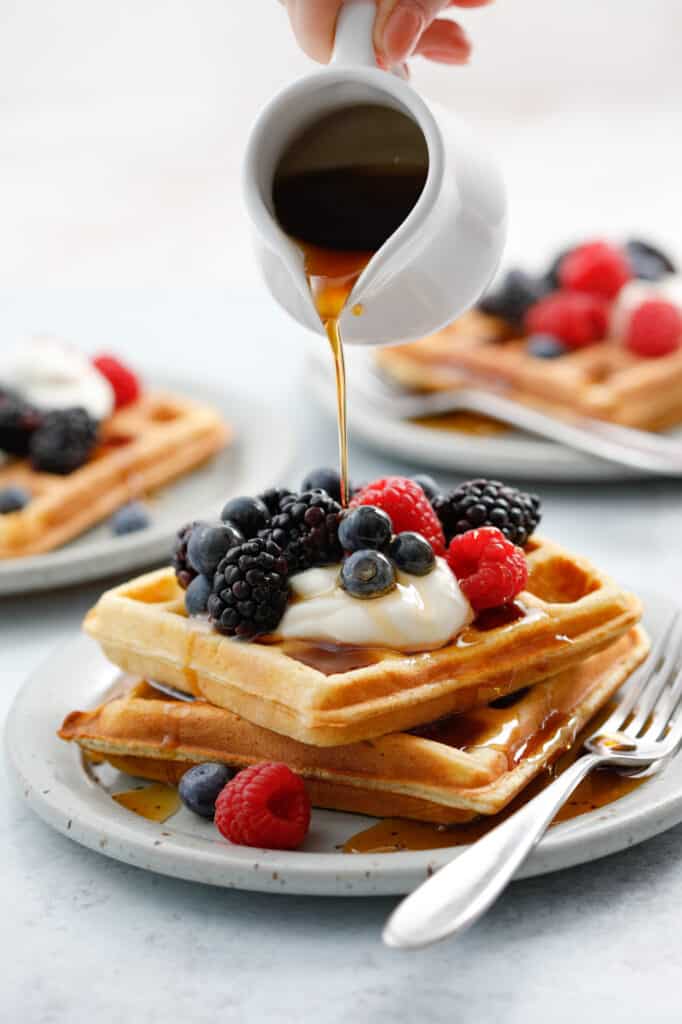 A stack of two waffles topped with sour cream and berries with maple syrup being drizzled on