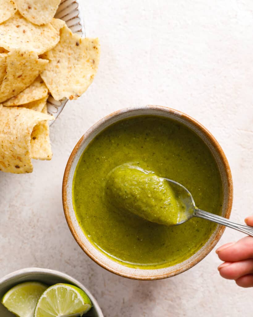 overhead view of a ceramic bowl with bright green salsa verde inside being spooned out with chips and limes in the background