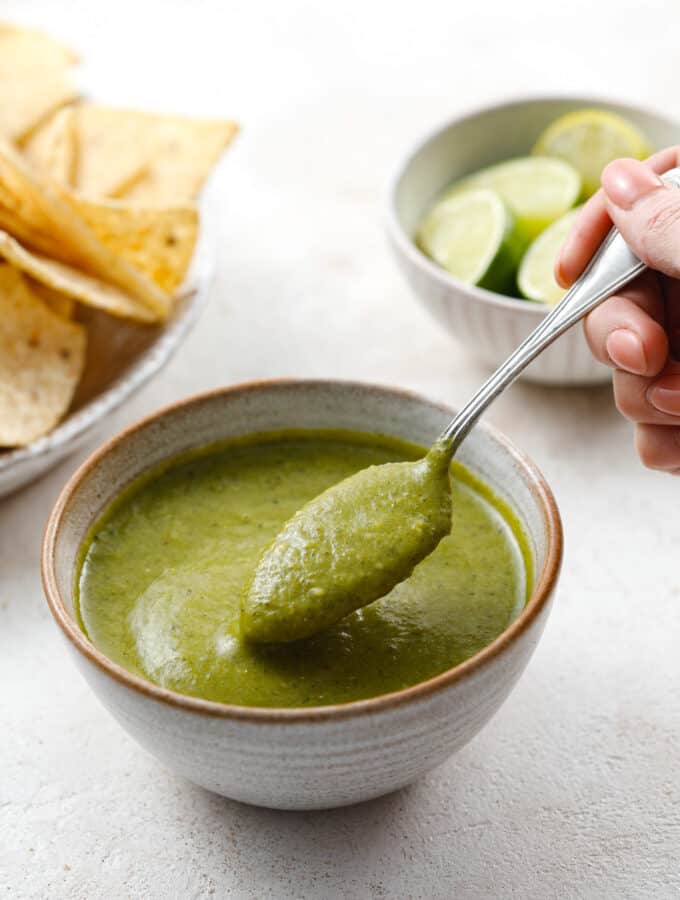 A bowl of bright green jalapeno salsa in a ceramic bowl being scooped with a teaspoon.