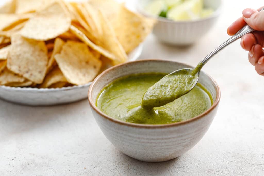 a ceramic bowl with bright green salsa verde inside being spooned out with chips and limes in the background