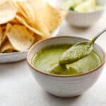 a ceramic bowl with bright green salsa verde inside being spooned out with chips and limes in the background