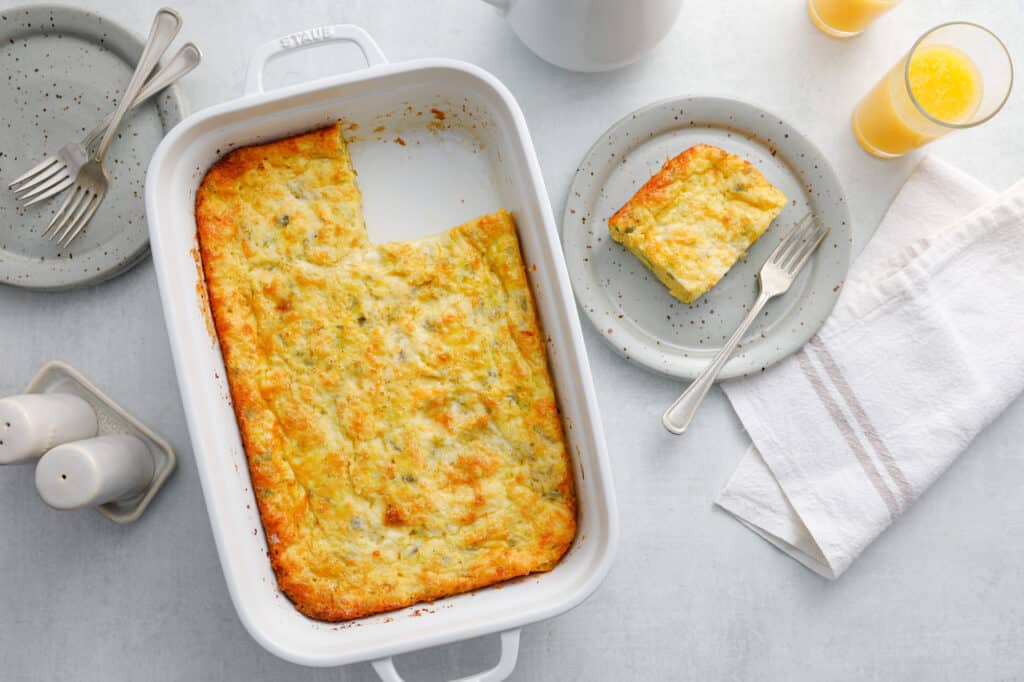 Slice of green chili egg casserole on a grey dish with a fork next to glasses of orange juice and a white napkin in a breakfast scene