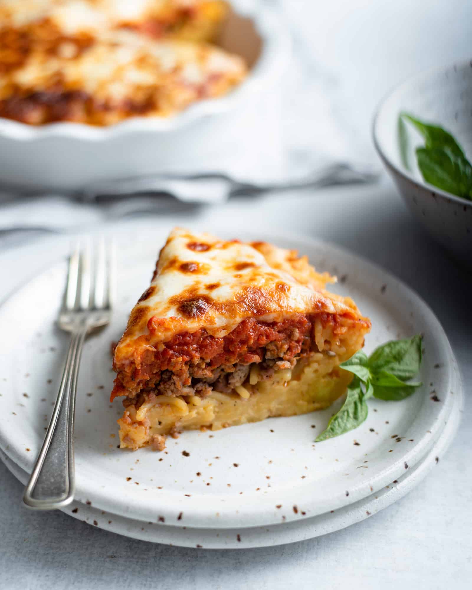 a slice of spaghetti pie on a plate with a fork, a spaghetti crust topped with ground Italian sausage, marinara and mozzarella