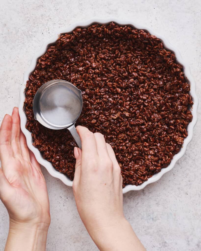 A chocolate rice Krispie crust being pressed into a pie plate with the back of a measuring cup