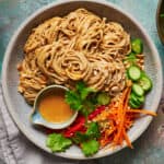 Overhead view of a bowl of peanut noodles with veggies and extra sauce on the side