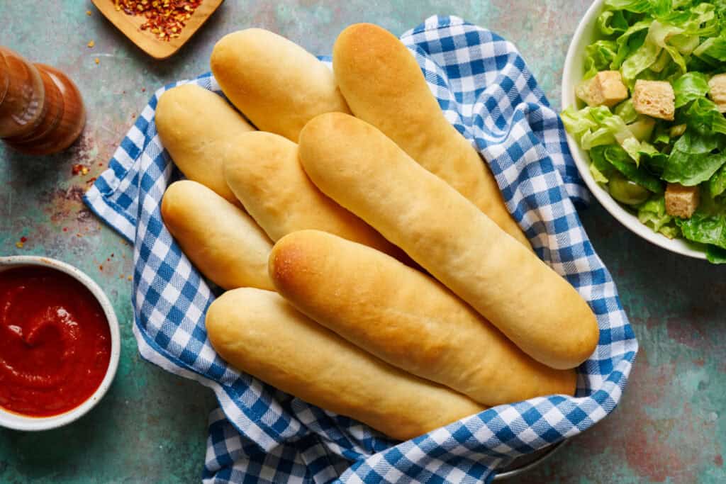 overhead view of breadsticks in a basket with gingham napkin with salad and marinara on the side.