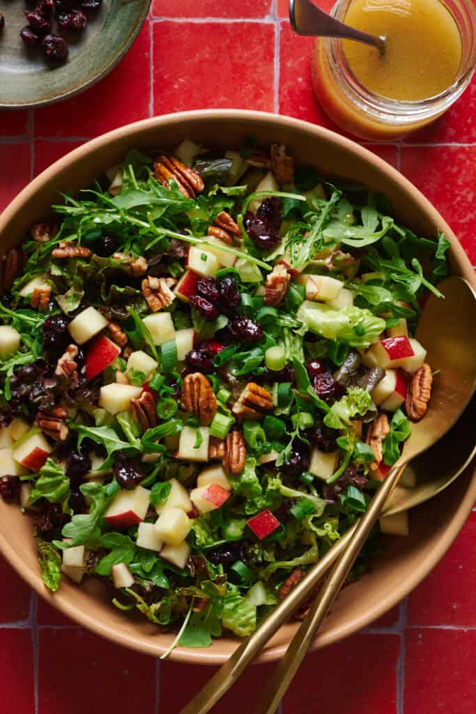 Arugula salad with apples, pecans, cranberries and scallions in a large salad bowl with tongs.