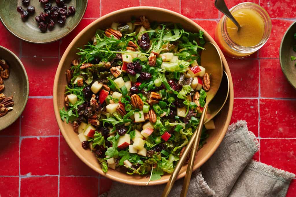 Arugula salad with apples, pecans, cranberries and scallions in a large salad bowl with tongs.