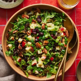 Arugula salad with apples, pecans, cranberries and scallions in a large salad bowl with tongs.