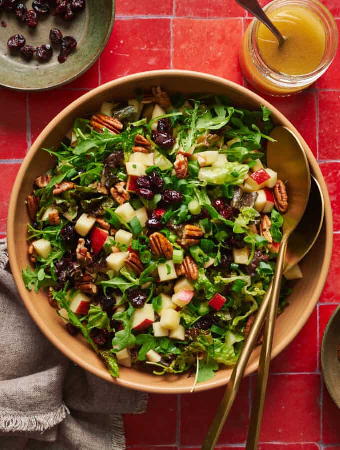 Arugula salad with apples, pecans, cranberries and scallions in a large salad bowl with tongs.