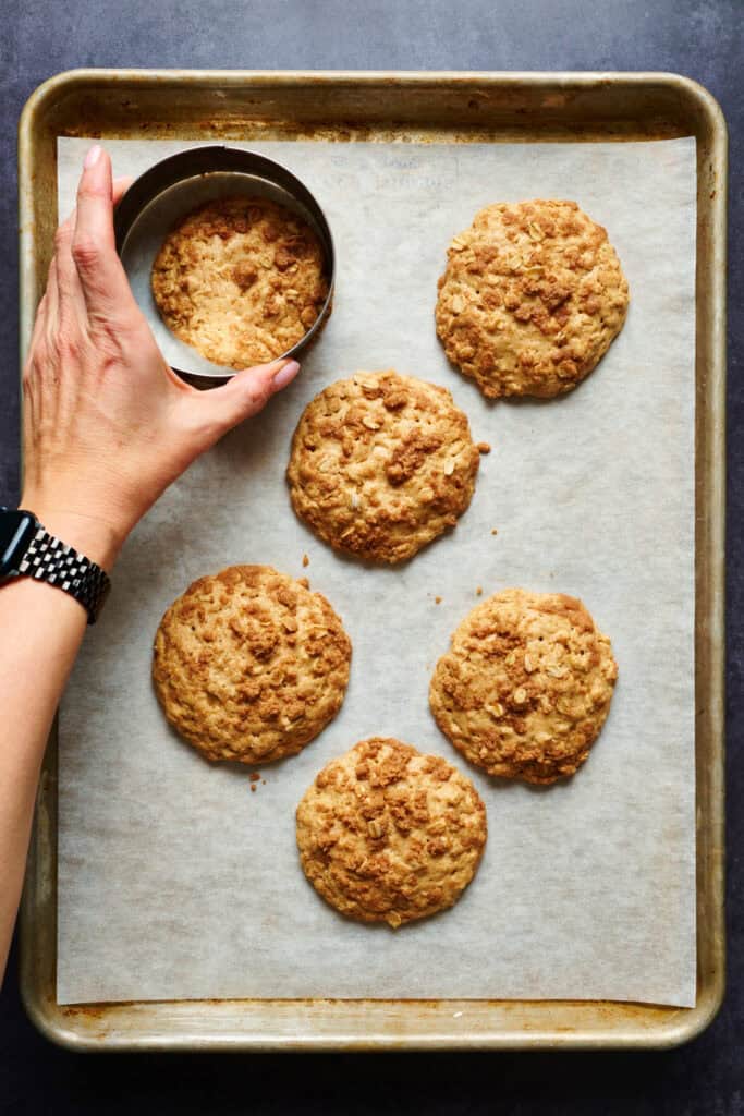 cookies baked on a sheet pan and a hand uses a cookie cutter to even out the edges