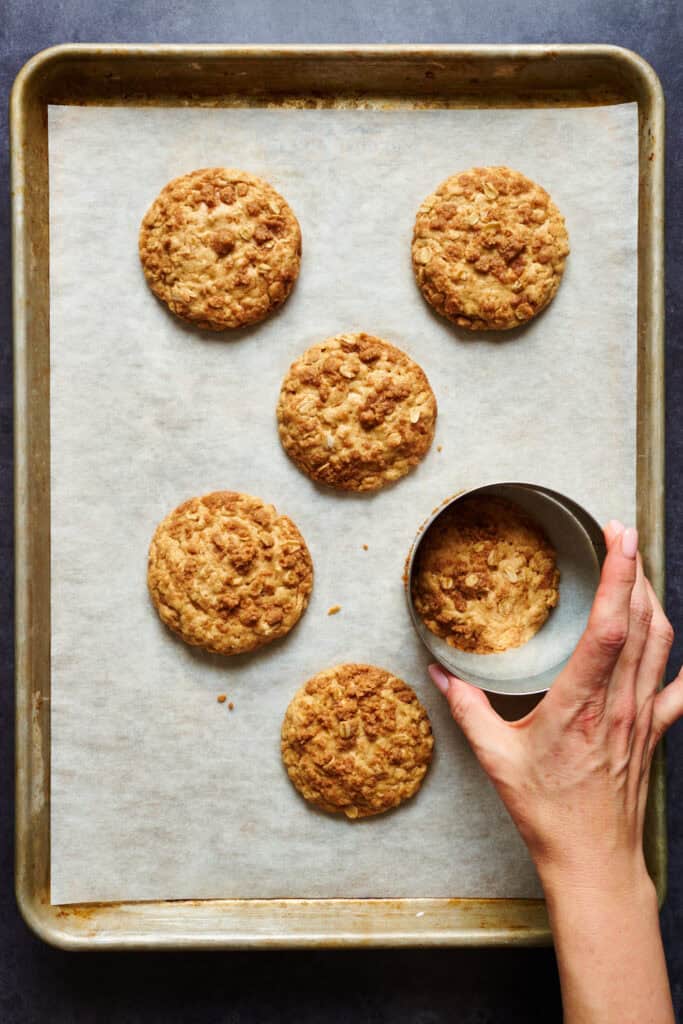 cookies baked on a sheet pan and a hand uses a cookie cutter to even out the edges having completed 5