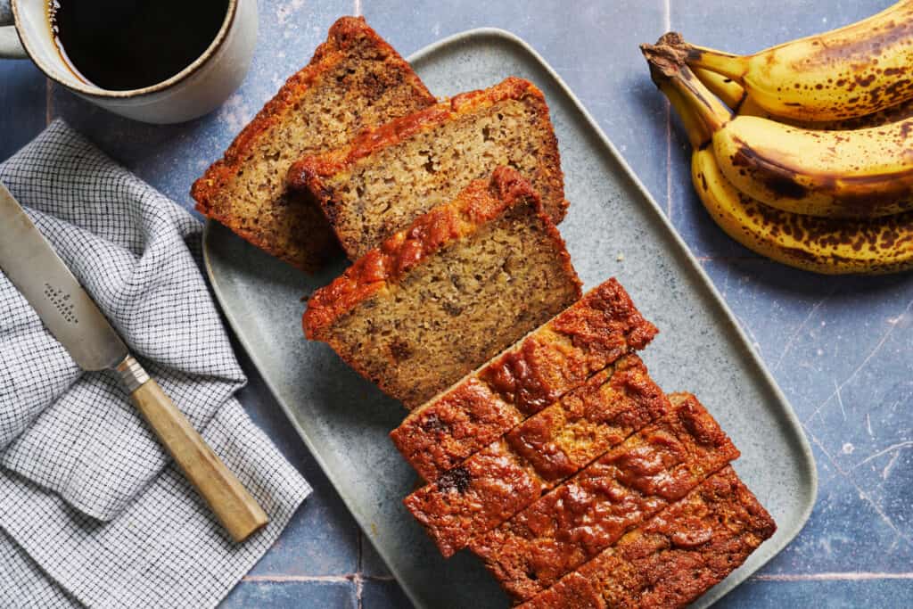 a loaf of sourdough discard banana bread, sliced with bananas and coffee on the sides