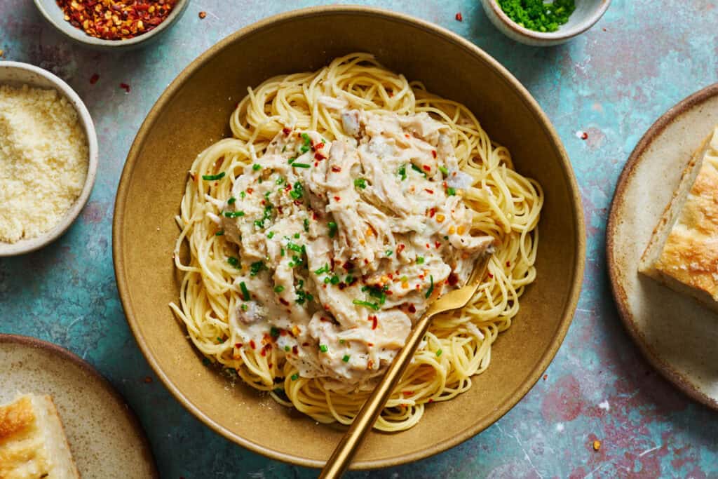 Shredded chicken in a white wine cream sauce over a bed of angel hair pasta in a bowl on a turquoise background.