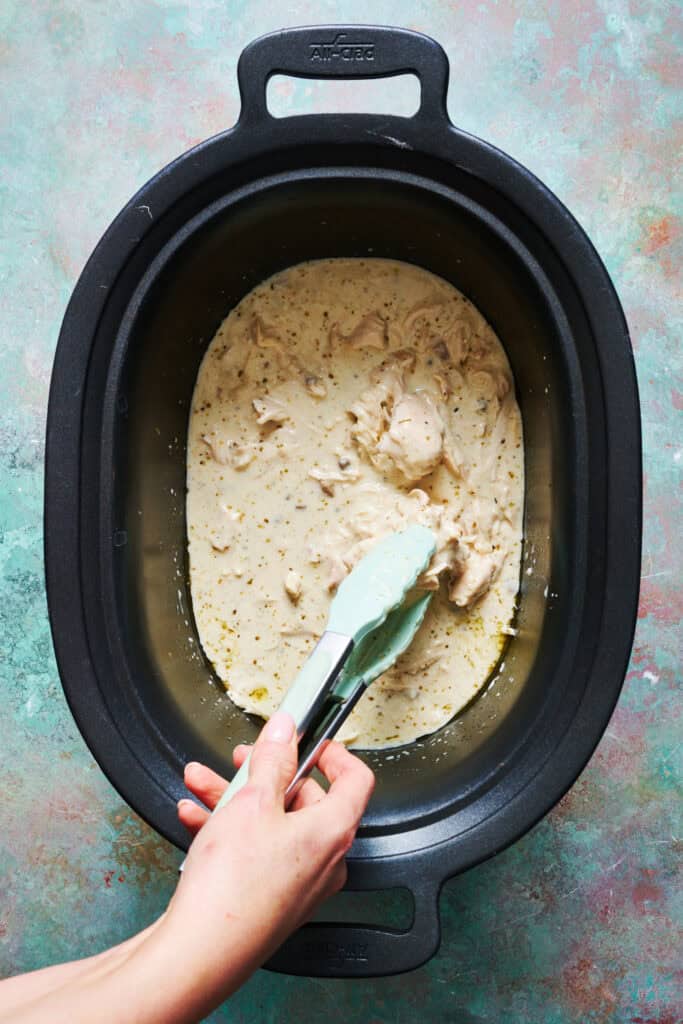 Chicken being broken up with tongs in a slow cooker with creamy sauce