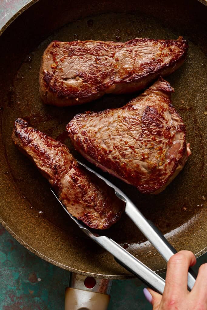 steak being seared in a pan with tongs