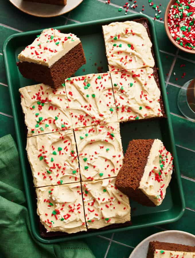 Gingerbread Cake with Cinnamon Molasses Frosting on a green sheet pan with a green napkin and tile background. Several slices removed from full cake.