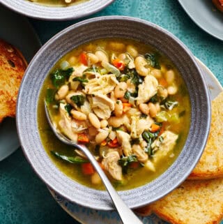 overhead view of a bowl filled with soup that has shredded turkey, beans, greens, broth, carrots and spinach. It has more bowls of soup surrounding and slices of bread on the side.