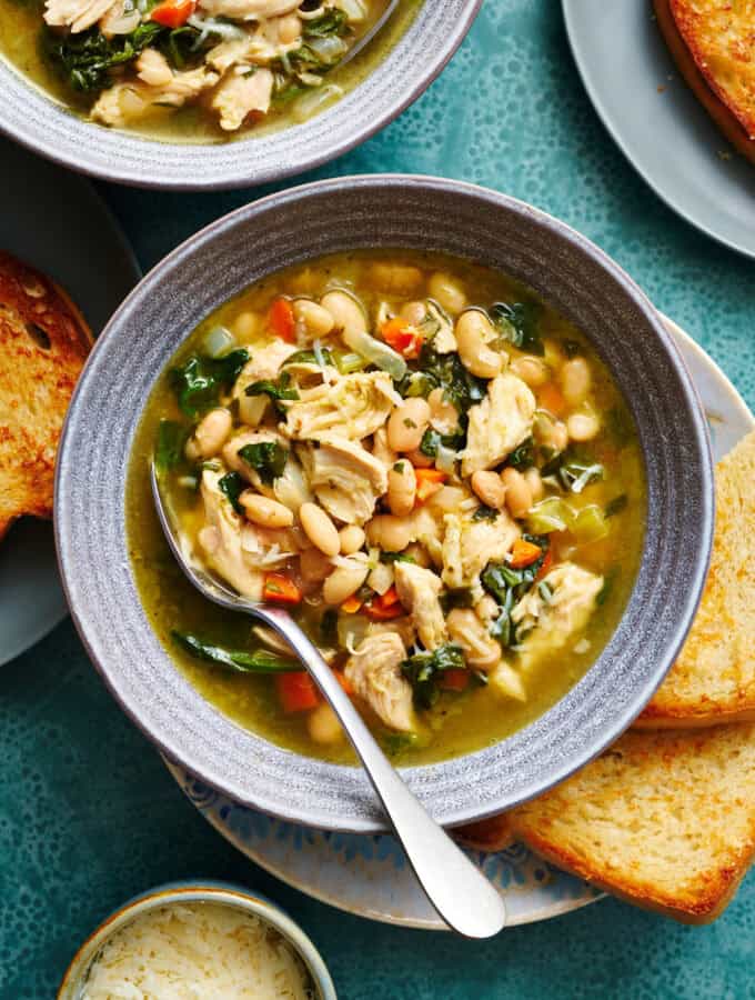 overhead view of a bowl filled with soup that has shredded turkey, beans, greens, broth, carrots and spinach. It has more bowls of soup surrounding and slices of bread on the side.