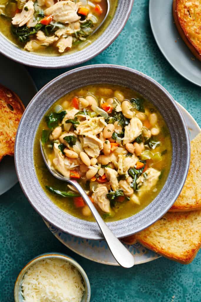 overhead view of a bowl filled with soup that has shredded turkey, beans, greens, broth, carrots and spinach. It has more bowls of soup surrounding and slices of bread on the side.