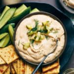 overhead view of a bowl of spicy dill pickle dip with veggies and crackers surrounding it as well as side plates with individual servings