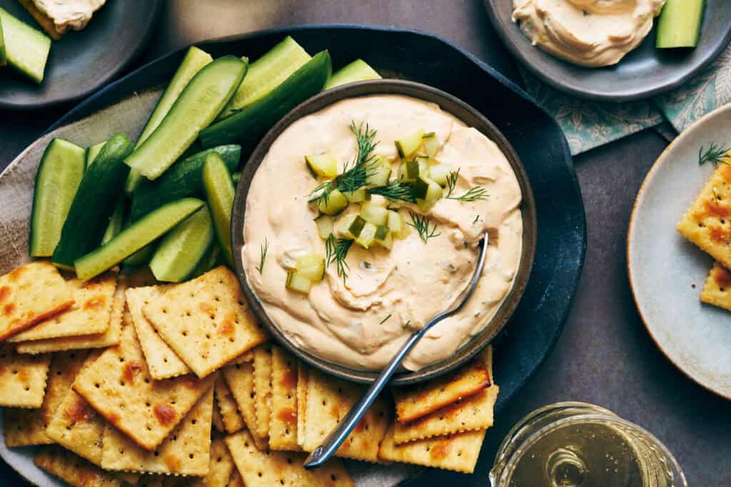 overhead view of a bowl of spicy dill pickle dip with veggies and crackers surrounding it as well as side plates with individual servings