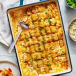 overhead view of a 9 x 13 pan filled with buffalo chicken tater tot casserole garnished with scallions and with a slice removed. Blue cheese and extra buffalo sauce are pictured on the side.