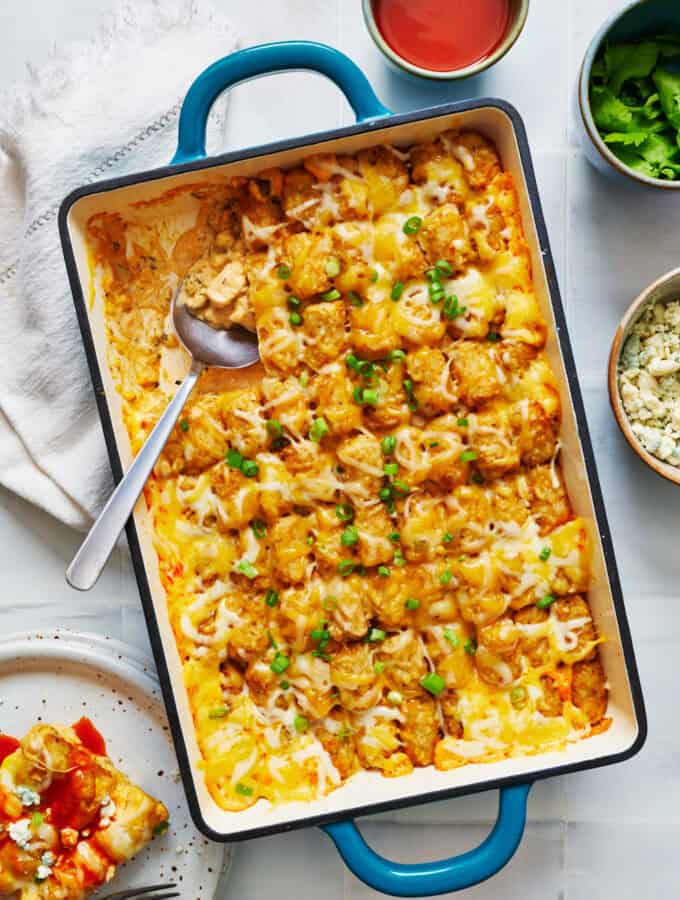 overhead view of a 9 x 13 pan filled with buffalo chicken tater tot casserole garnished with scallions and with a slice removed. Blue cheese and extra buffalo sauce are pictured on the side.