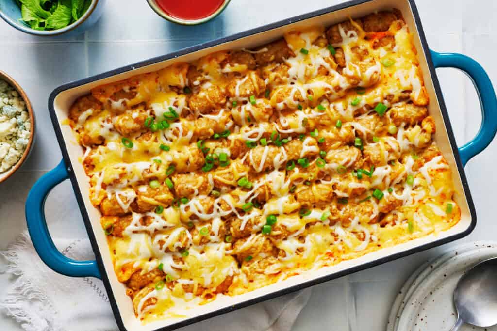 Overhead view of a casserole dish filled with tater tots casserole with buffalo chicken and buffalo sauce and blue cheese on the side.